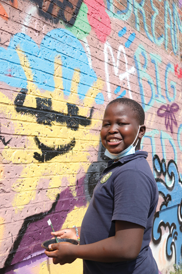 A child stands in front of a wall graffitied with colourful drawings.