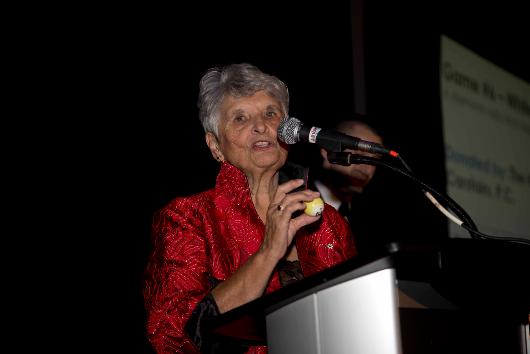 An older woman wearing a bright red blazer speaks into a microphone at a podium.